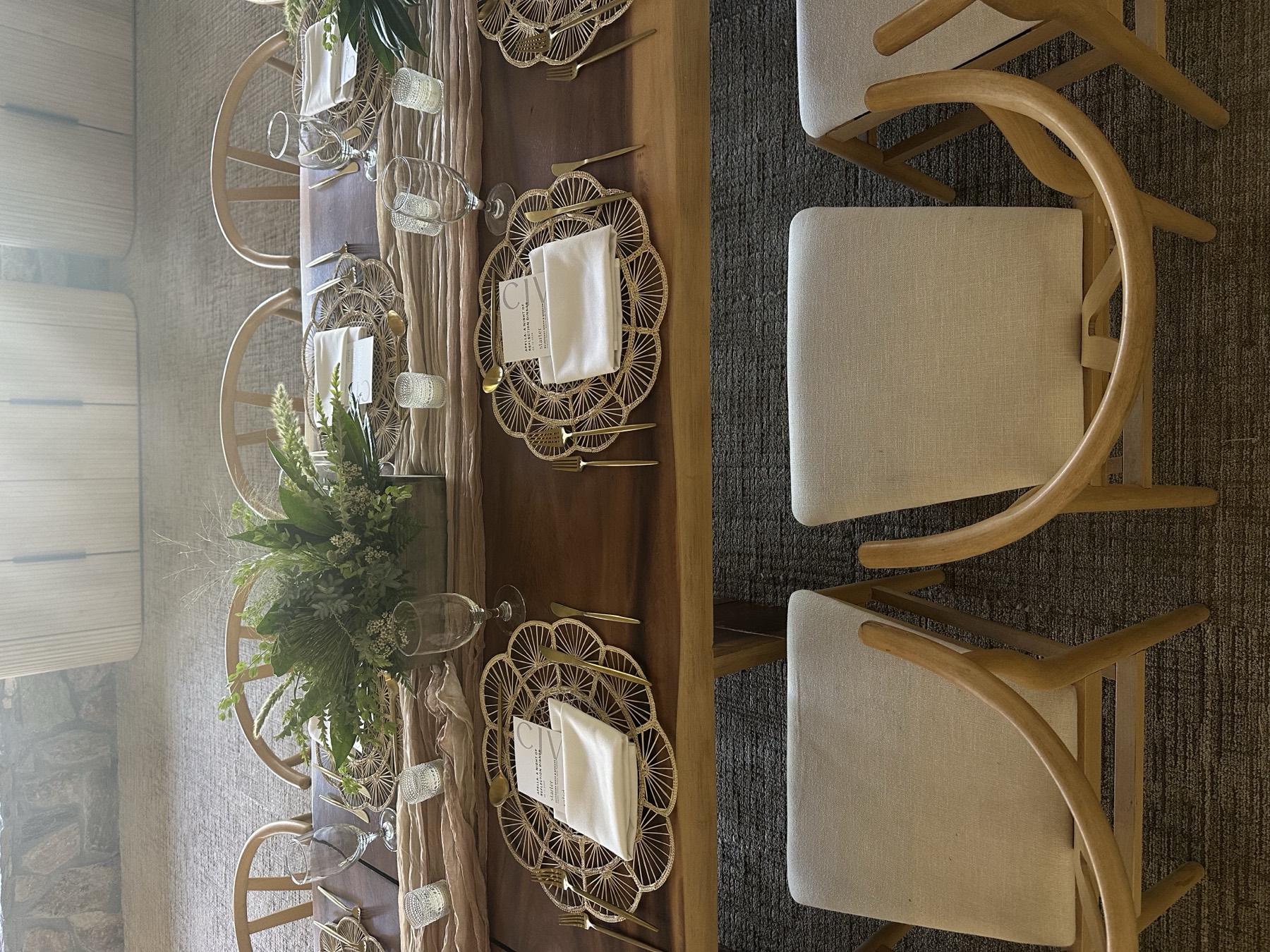 An overhead view of a long wooden table set with woven chargers, gold flatware, greenery and printed menus