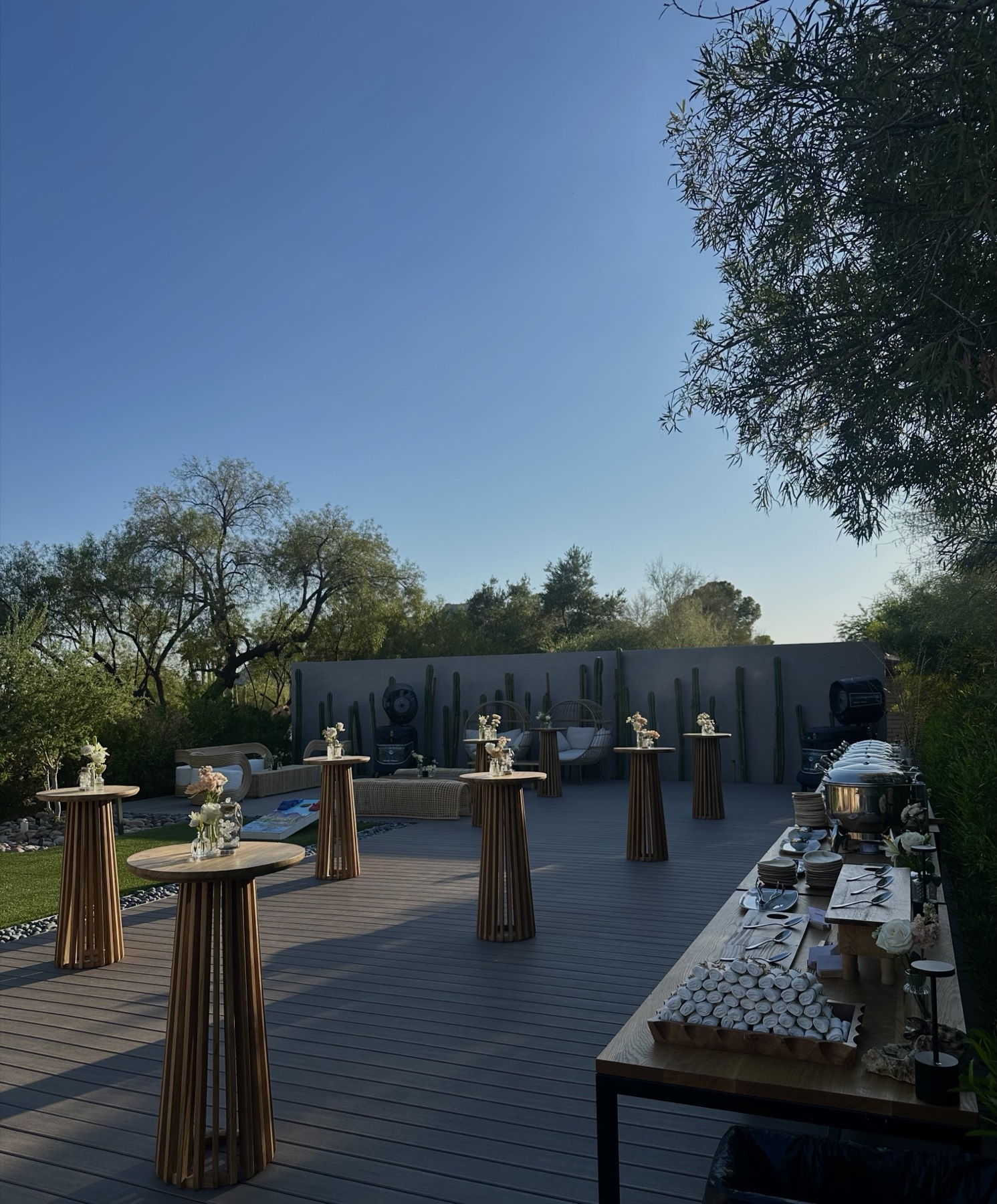 Cocktail tables and a buffet line set out on a deck at dusk, desert planting and a long wall behind